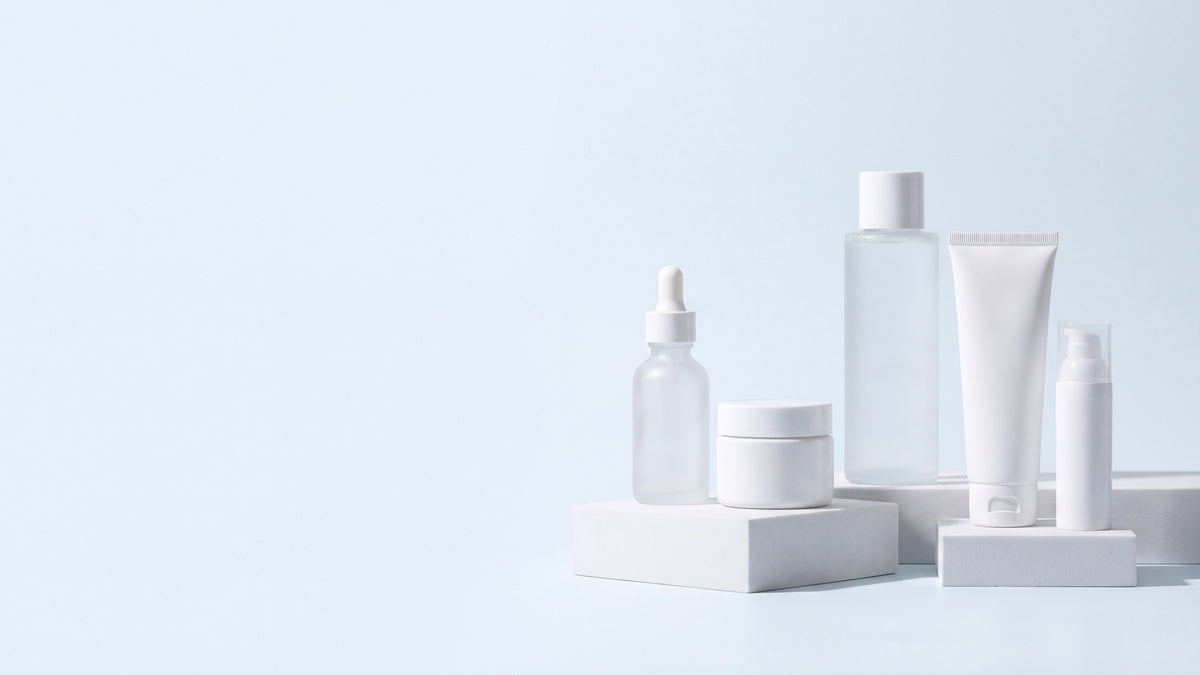 Unlabeled skincare bottles and jars on white plinths against a pale blue backdrop